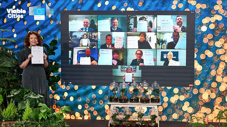 Bildskärm där alla signerande parter syns tillsammans i ett videosamtal. Bildskärmen är placerad på en scen dekorerad med växter.