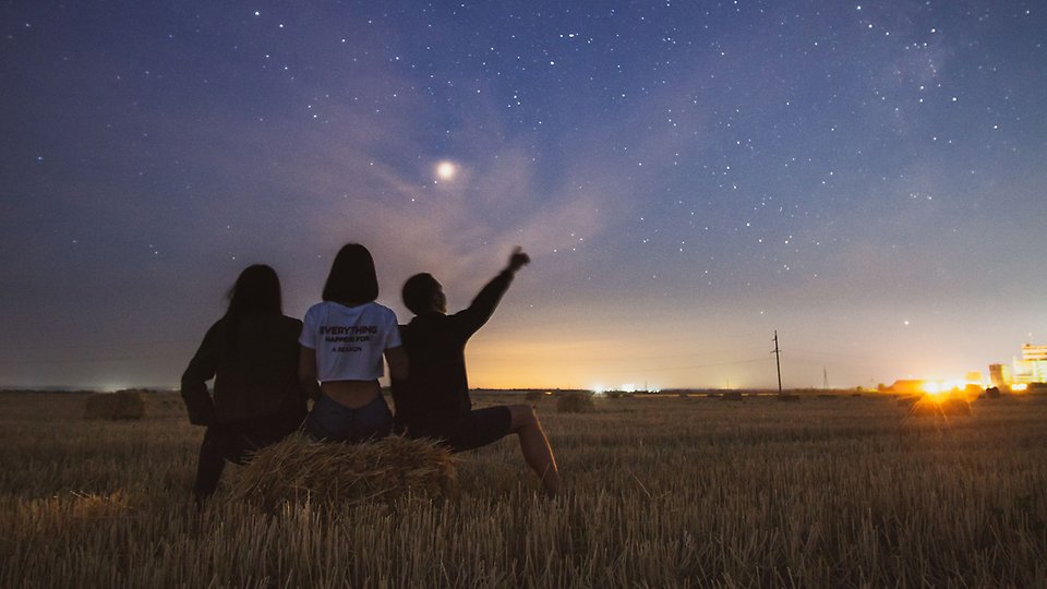 Tre personer sitter på en höbal och blickar upp mot en stjärnklar himmel.