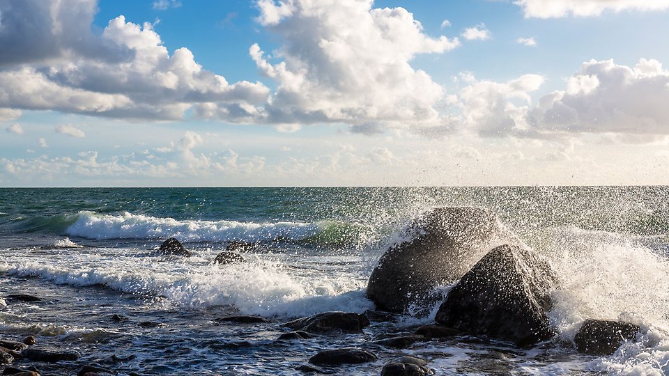 Vågor på havet en solig dag. Vågorna slår mot två stora stenar och vattnet skvätter över dem.